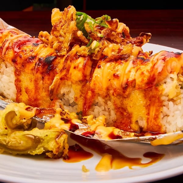 A large sushi roll with white rice, topped with orange sauce, fried toppings, and green onions.