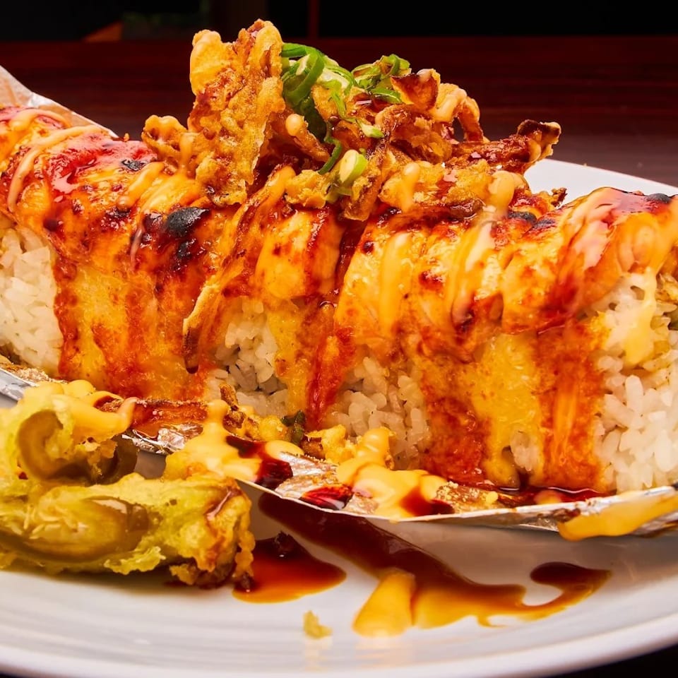 A large sushi roll with white rice, topped with orange sauce, fried toppings, and green onions.