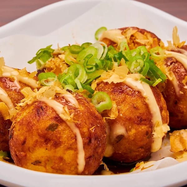 Four golden-brown takoyaki balls topped with mayonnaise, bonito flakes, and green onions in a white dish.